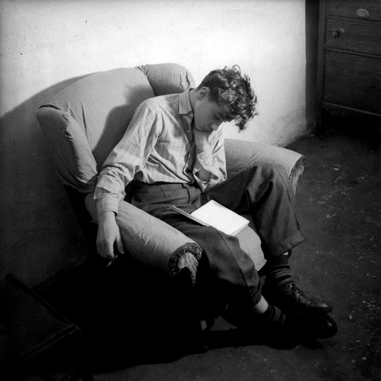 a black and white photo of richard soakup - a young white man with short curly brown hair - sitting in an armchair with his head rested on his right hand, seemingly asleep, with an open book in his lap and cigarette in his left hand that is hanging down from the armchair. he is dressed in a white pinstriped button up shirt with 2 buttons undone, grey pleated dress pants, warm socks and black dress shoes